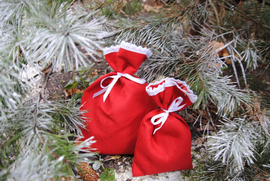 Two small red  linen gift bags with white lace trim and white ribbons, placed among frosty pine branches outdoors.