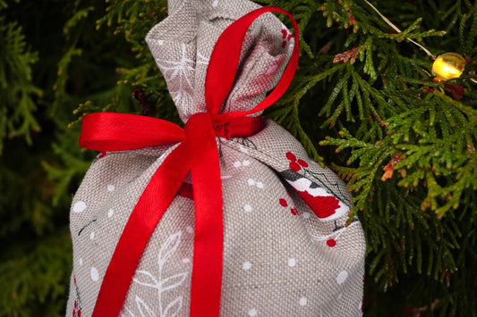 A small linen  fabric gift bag tied with a bright red ribbon bow, hanging among green pine branches with a warm string light in the background.
