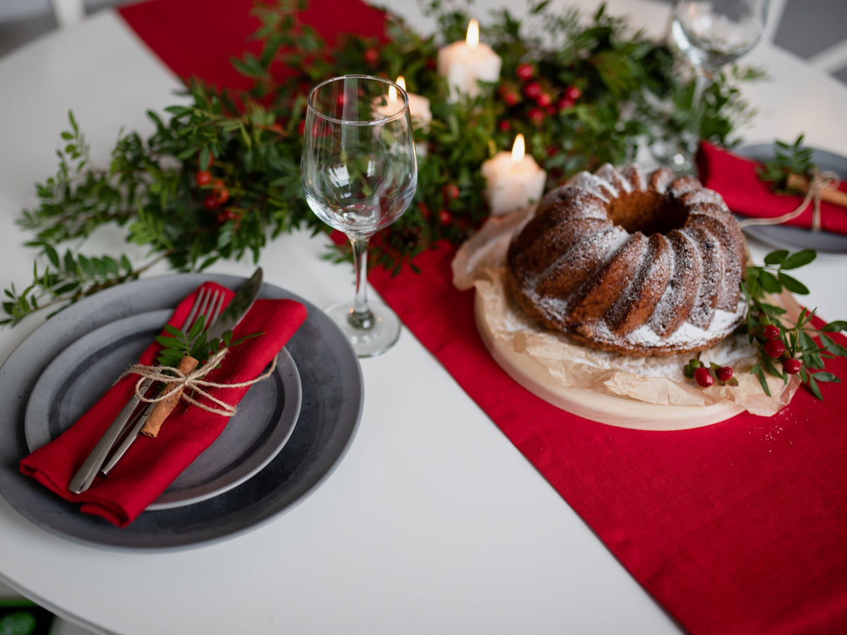 A holiday-themed table setting featuring a ring-shaped bundt cake dusted with powdered sugar on a wooden board, surrounded by green garlands, red candles, and a place setting with a red linen napkin and gray plates on a red linen table runner.