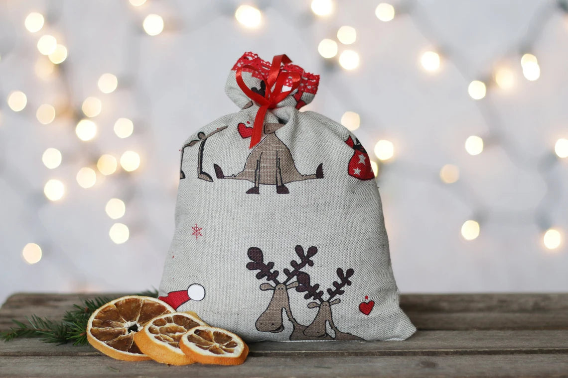 A festive linen Christmas sack with a red ribbon bow, decorated with illustrations of Santa Claus and reindeer, sitting on a wooden surface with dried orange slices and greenery against a warm bokeh background.