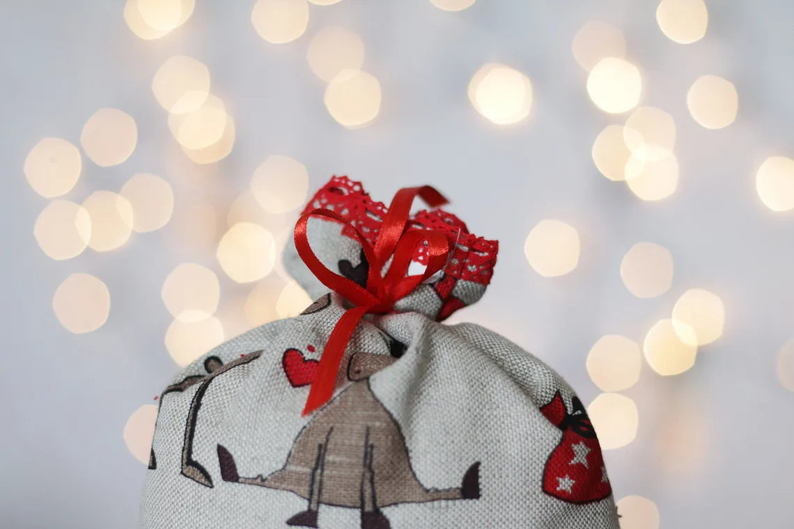 A linen fabric gift pouch with a rustic, holiday-themed print featuring a reindeer, a red heart, and a red ribbon bow, set against a soft-focus background of warm bokeh lights.