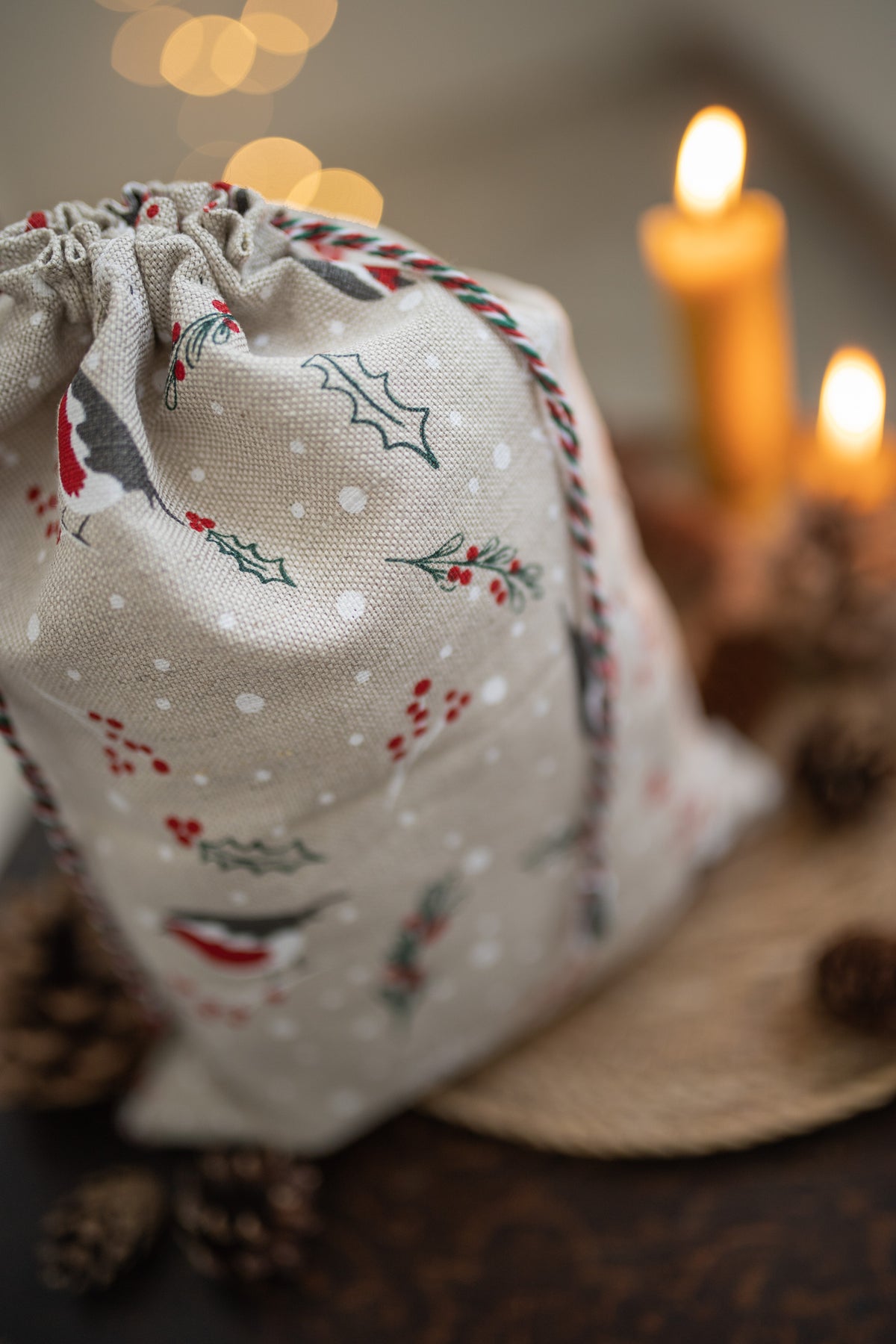 A beige fabric drawstring gift bag with a Christmas print of holly, mistletoe, snowflakes, and robin , styled on a dark wooden surface with lit candles and pinecones in the background.