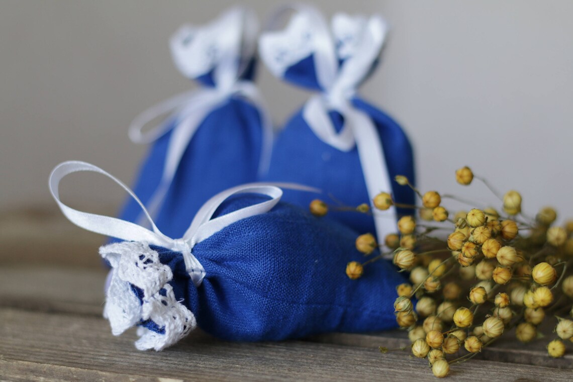 Three small royal blue linen pouches tied with white ribbons and decorated with white lace trim, arranged on a rustic wooden surface next to dried yellow flowers.