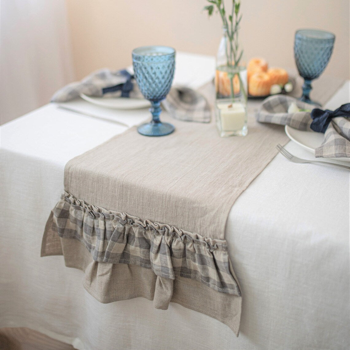 A beige linen table runner with a ruffled plaid trim, styled on a white tablecloth set dining tables with blue glasses, plates, and napkins.