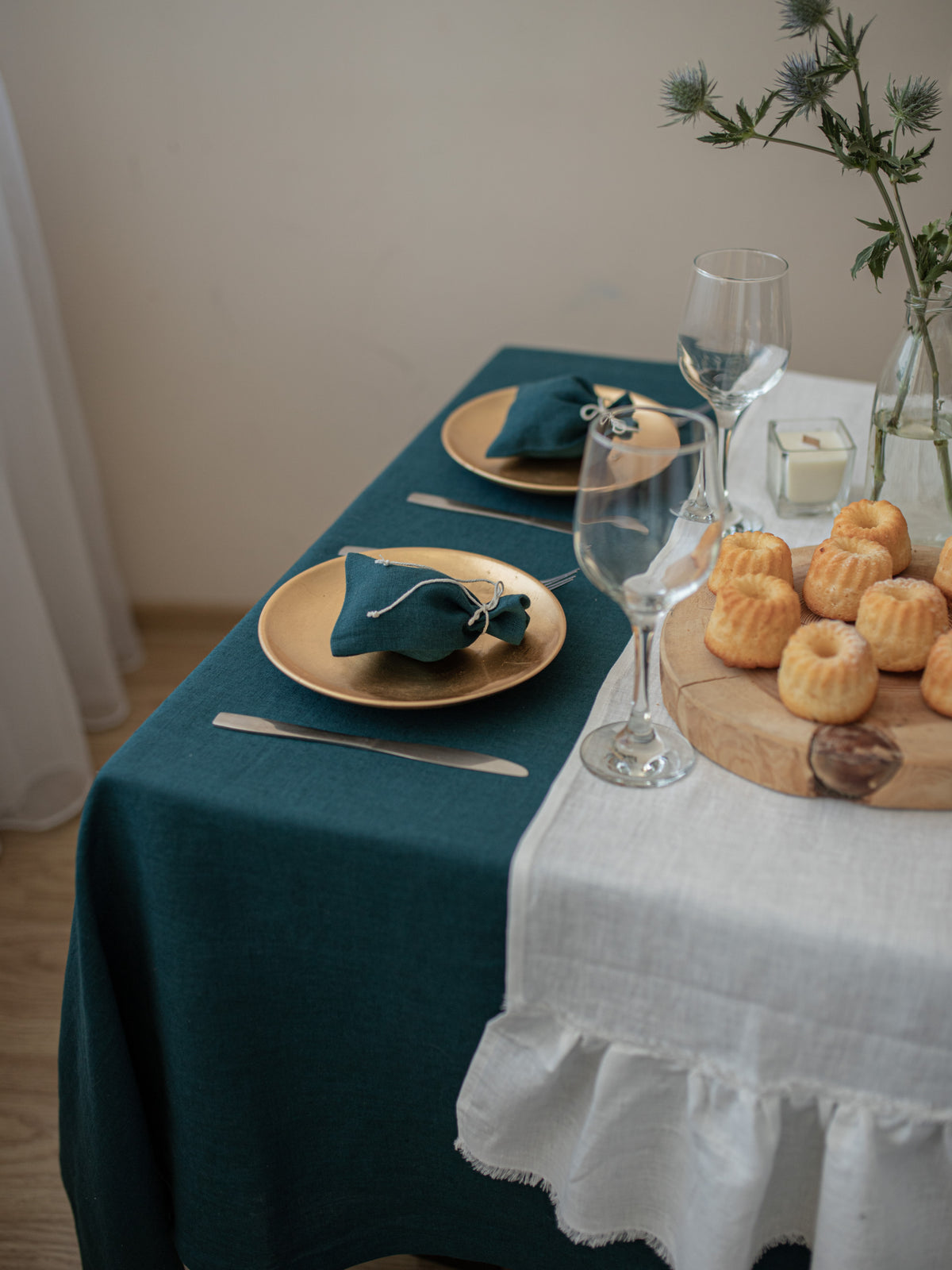 Dining table set with teal tablecloth, off white linen table runner plates, glasses, and a wooden board with pastries.