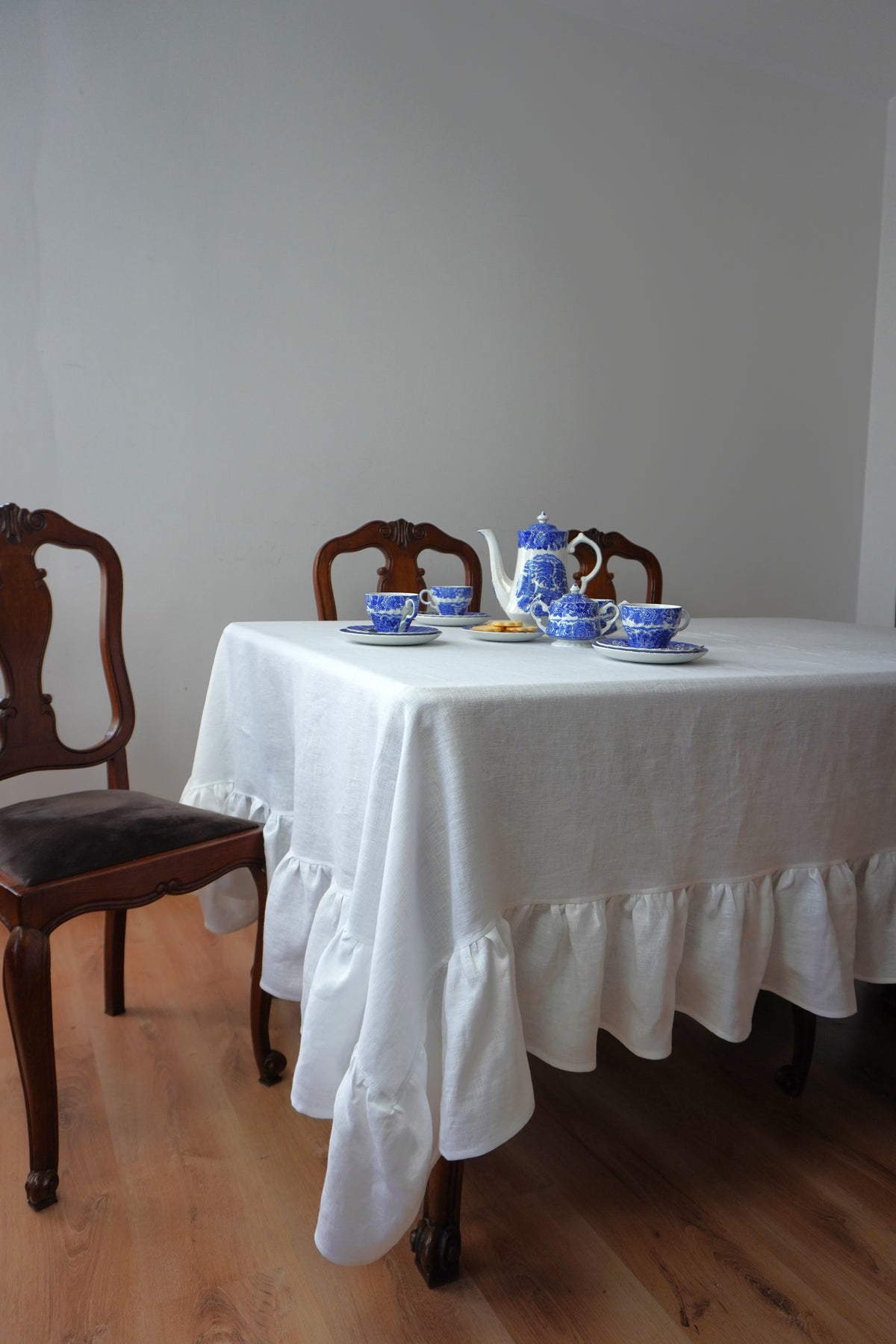 Dining table set with a white linen  tablecloth, blue teacup, and teapot in a room with wooden floor and white walls.