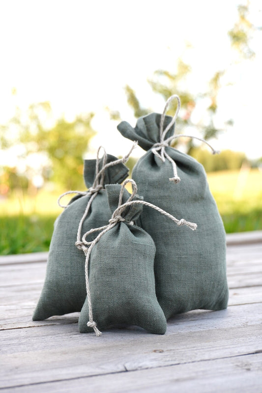 Three small sage green linen pouches of different sizes arranged on a weathered wooden deck with a blurred outdoor garden background.