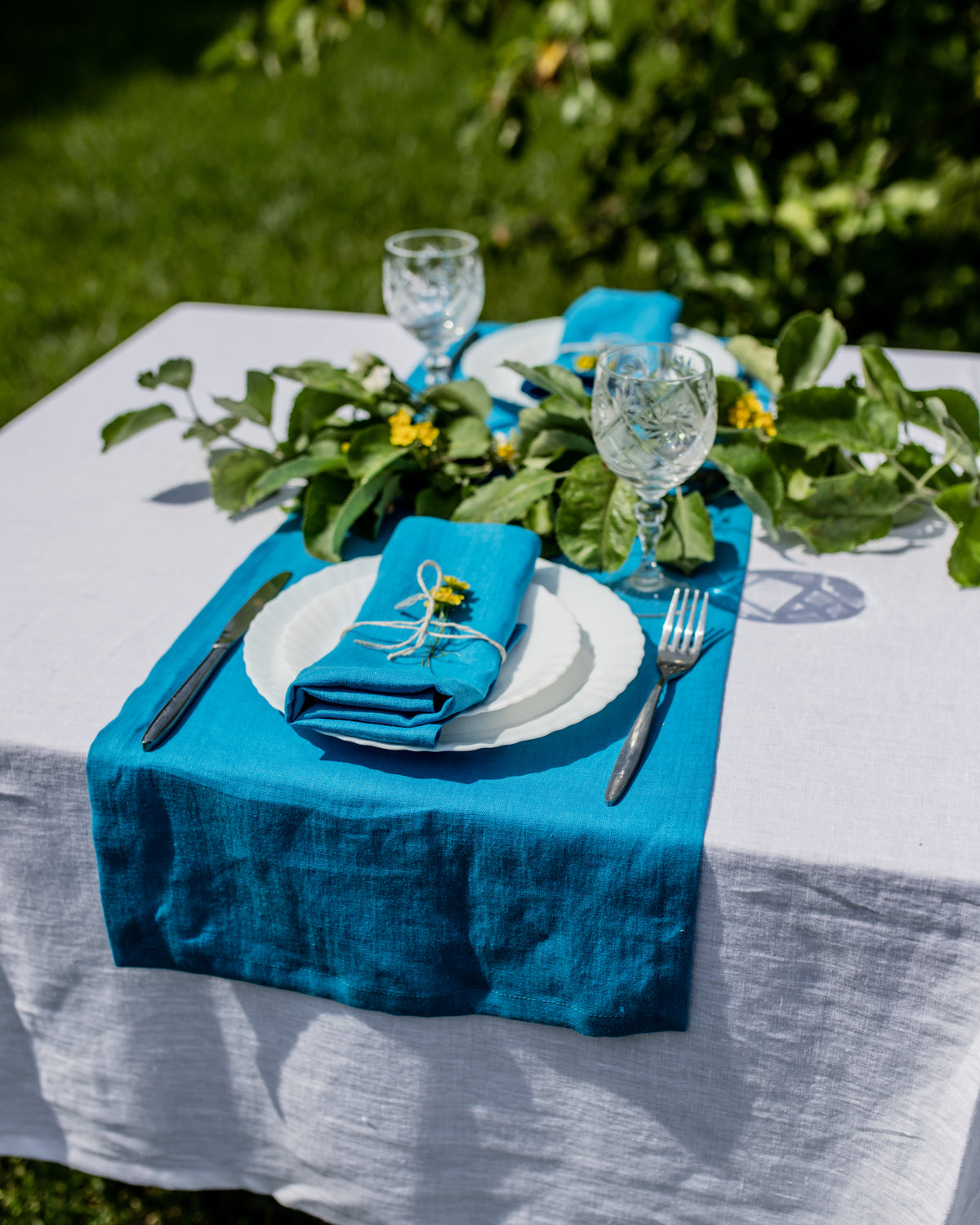 Table setting with blue table runner, white plates, and glasses on a white tablecloth outdoors.