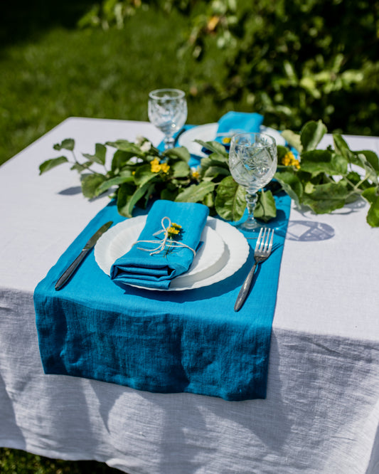 Table setting with blue table runner, white plates, and glasses on a white tablecloth outdoors.