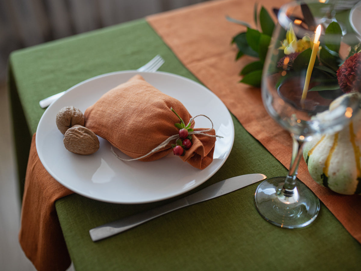 An autumn-themed table setting featuring a white plate with a burnt orange linen napkin tied with twine and a small sprig of red berries, accompanied by walnuts, a wine glass, and a candle on a rustic table runner.