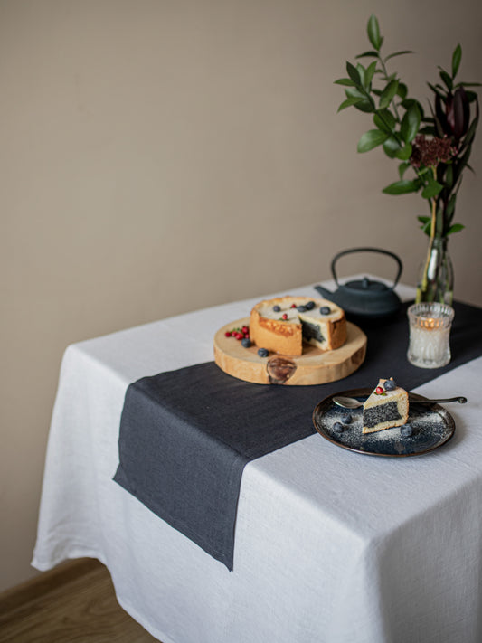 Table setting with a cake, teapot, and plate of dessert on a white linen tablecloth.