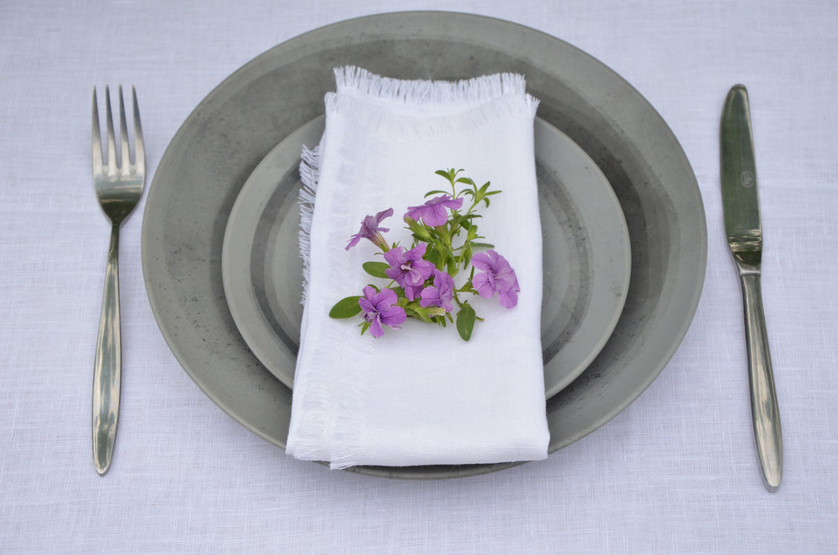 Table setting with gray plates, white fringed linen napkin with purple flowers, fork, and knife on a white tablecloth.