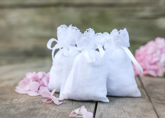 Three small white linen fabric gift bags with lace trim and white ribbon ties, arranged on a rustic wooden surface with pink flower petals around them.