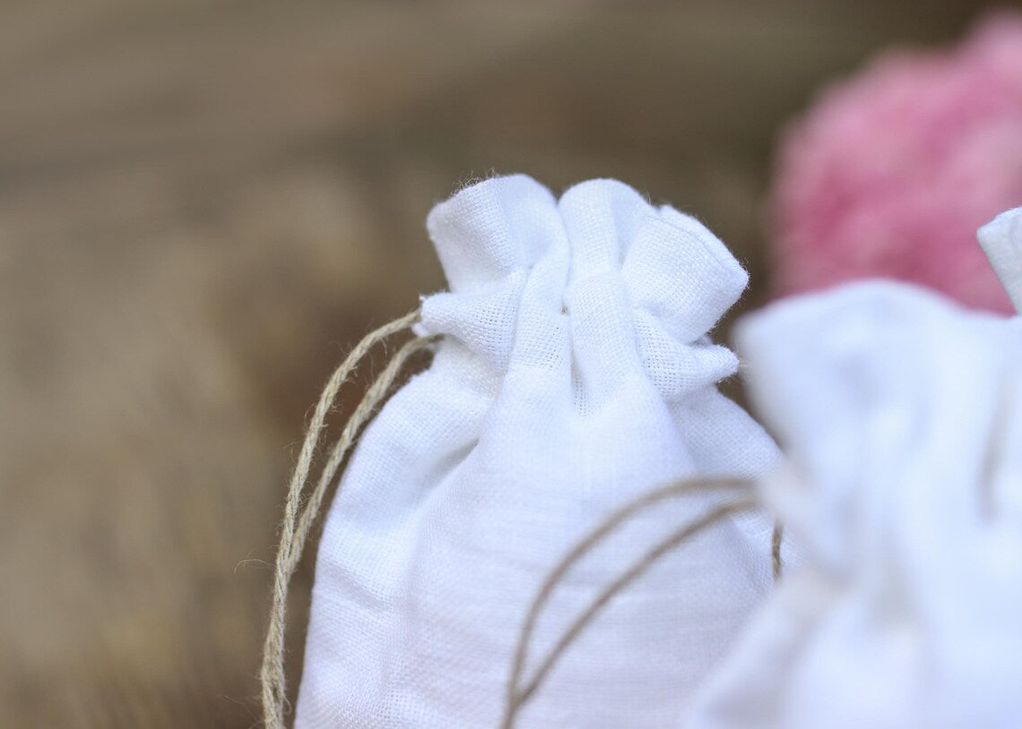 A close-up of a small white linen drawstring pouch with a natural jute twine cord, set against a soft-focus warm brown background 