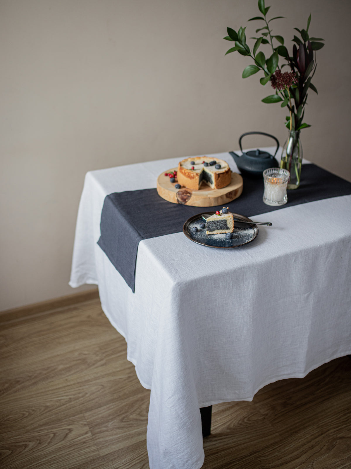 Table setting with a white linen tablecloth, charcoal gray runner, and food items on a wooden floor.