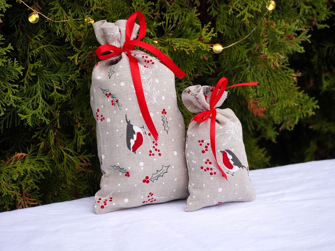Two small fabric gift bags with a bird and holly berry print on a light beige background, tied with bright red ribbons, set against a green evergreen tree with warm string lights.