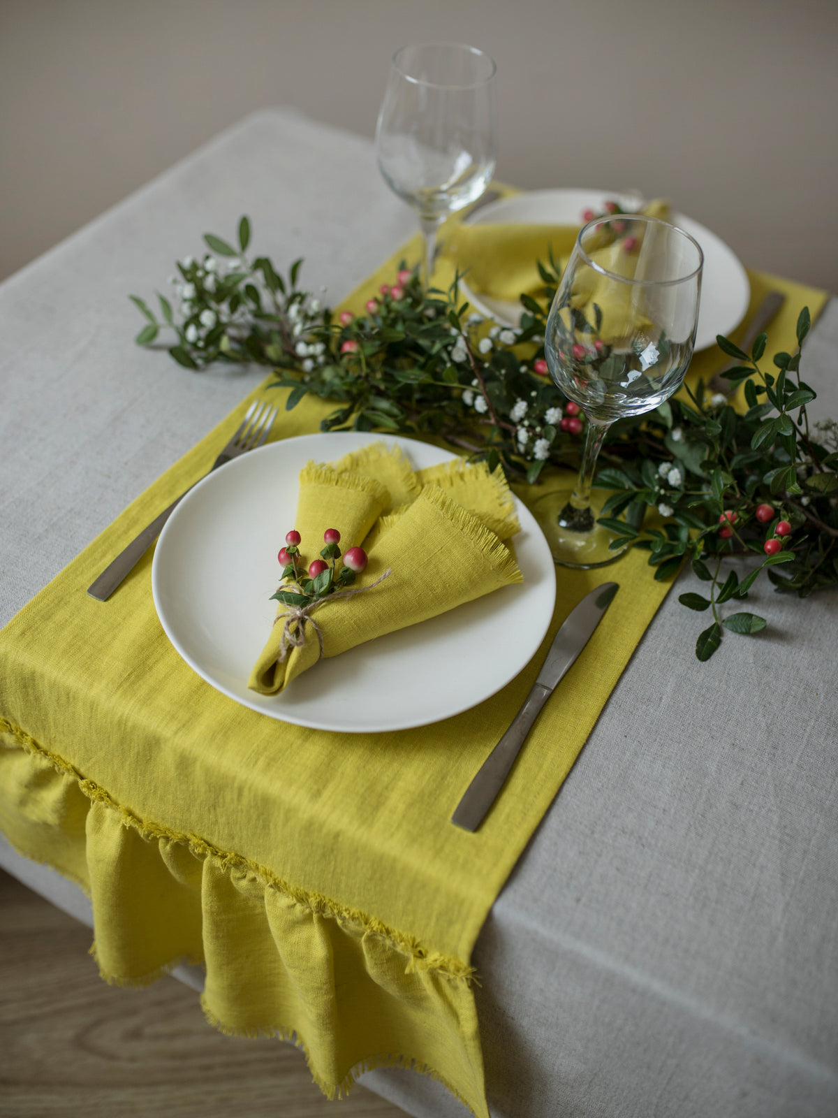 A styled dining table setting featuring a bright yellow (chartreuse)  linen table runner with a ruffled hem, matching yellow  (chartreuse) napkins on white plates, silver flatware, and green berry garlands.