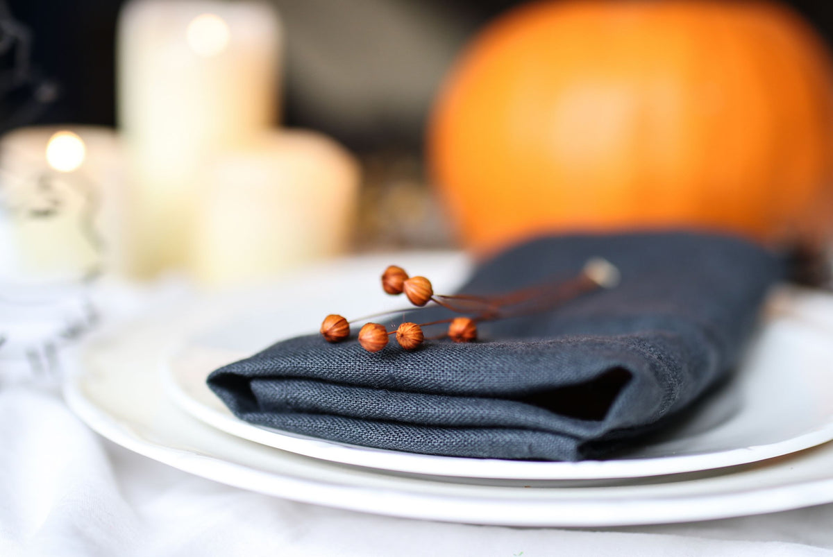 Black linen napkin placed on a white plate with a blurred background featuring a pumpkin, suggesting a Halloween theme.