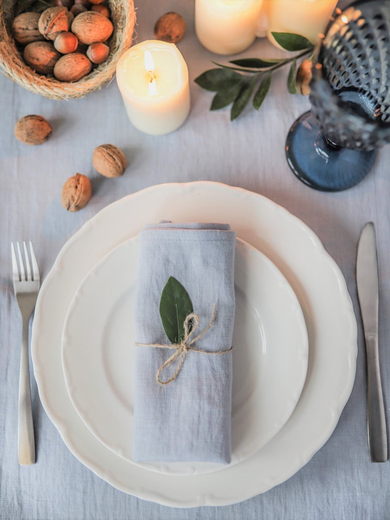 A dusty blue linen napkin is placed on a white plate, tied with a ribbon, presented on a table with a white tablecloth, candles, and decorative elements.