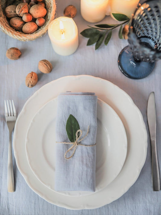 A dusty blue linen napkin is placed on a white plate, tied with a ribbon, presented on a table with a white tablecloth, candles, and decorative elements.